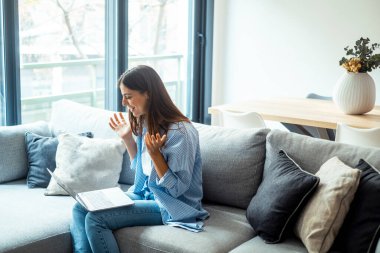 Genç kadın el sallıyor. Gülümseyen kadın dizüstü bilgisayar kullanıyor, evdeki koltukta oturuyor, güzel kız sosyal ağlarda sohbet ediyor ve arkadaşlarıyla gülüyor. Teknoloji ve arkadaşlık kavramı..