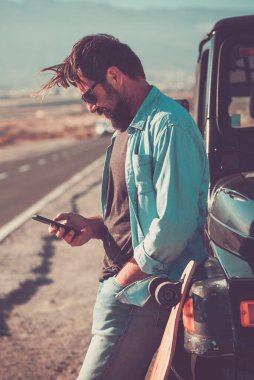  Adam gezgin, arka planda uzun manzaralı düz bir yol ile arabanın dışında telefon ve gezinti bağlantısı kullanıyor. Macera ve modern yaşam tarzı insanları. Yetişkinler bir araca karşı açık havada rahatlar.