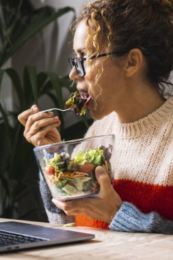 Ev ödevi yapan, salata yiyen ve masada bilgisayar başında çalışan modern iş kadınının portresi. Öğle tatili ve iş yoğunları. Diyet yapan sağlıklı bir kadın sebze yer.
