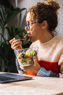 Çağdaş girişimci yeni iş konsepti konsepti yaşam tarzı iş sırasında yemek yemek, öğle yemeği zamanında. Dizüstü bilgisayar kullanan ve şeffaf kaseden salata yiyen bir kadın. Girişimci sağlıklı gıda yaşam tarzı