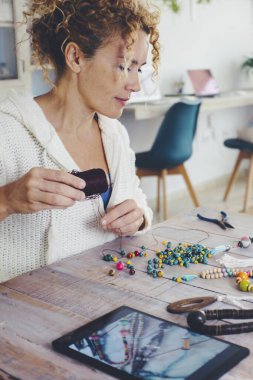 Boş zaman aktivitesi konsepti olan insanlar. Oturma odasındaki masada üzerinde tablet cihazıyla boncuk kolye yapan yetişkin bir kadın. Modern alternatif yeni iş çalışmaları İnternet 'te satılıyor