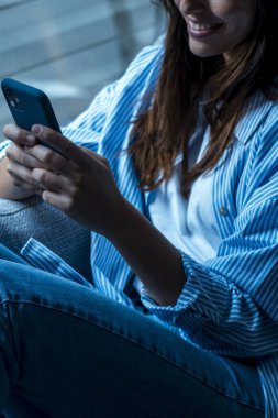 Milenyum boyunca rahat koltukta rahat rahat uzanan akıllı telefonu elinde tutan mutlu bir kadın. Gülümseyen genç bayan sosyal ağlarda sohbet ediyor, komik videolar izliyor, evde mobil uygulamalar kullanıyor..