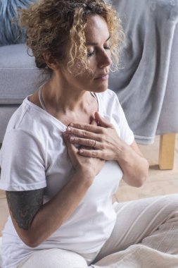 Orta yaşlı bir kadın evde meditasyon yapıyor oturma odasında halının üzerinde oturuyor gözleri kapalı ve lotus yogası pozisyonu almış. Nefes alma ve rahatlama rutini. Sağlıklı yaşam tarzı, kapalı alandaki kadınlar.