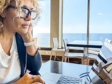 Gemi gezisi sırasında masada oturan güzel sarışın yönetici. Kıvırcık saçlı ve siyah gözlüklü bir kadın seyahat ederken laptop kullanıyor. Dijital göçebeler kavramı.