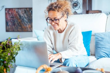 Mutlu kadın gülümsüyor ve dizüstü bilgisayarda yazıyor. Evde, kanepede rahat rahat oturuyor ve günlük giysiler giyiyor. Yeni işletme akıllı çalışma konsepti hayat tarzı evden işe gidiyor. Bilgisayar ağı kullanan neşeli bayan