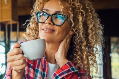 Serene woman at home drinking tea or coffee and enjoying relax leisure indoor activity alone sitting at the table. Portrait of cheerful and happy female people smiling and wearing eyewear. Curly hair