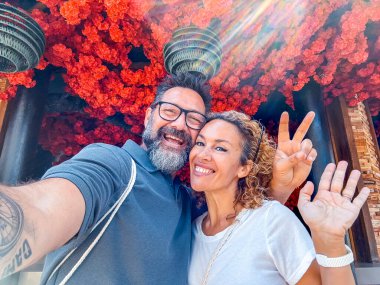 Mutlu orta yaşlı çift arka planda kırmızı çiçeklerle selfie çekiliyor. Seyahat yaşam tarzında turistik tatil aktivitelerinin keyfini çıkarıyorlar. Neşeli erkek ve kadın çevrimiçi içeriği paylaşıyor