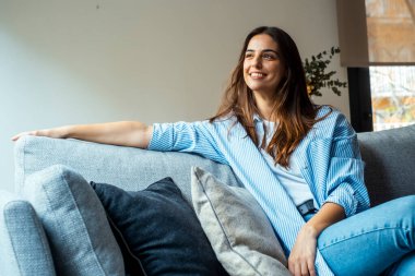 Rahatla ve gevşe. Genç, güzel, esmer, mutlu bir yüzü olan kadın kanepede tek başına gülümsüyor. Evde dinlenen rahat kadın portresi..
