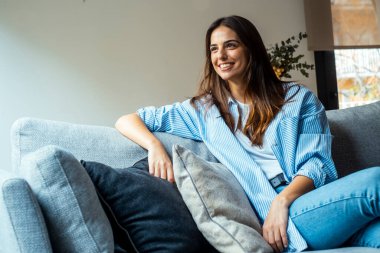 Rahatla ve gevşe. Genç, güzel, esmer, mutlu bir yüzü olan kadın kanepede tek başına gülümsüyor. Evde dinlenen rahat kadın portresi..