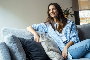 Rahatla ve gevşe. Genç, güzel, esmer, mutlu bir yüzü olan kadın kanepede tek başına gülümsüyor. Evde dinlenen rahat kadın portresi..