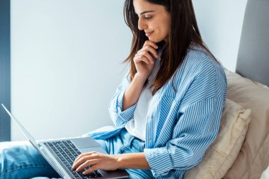 Genç bir kadın dizüstü bilgisayarda çalışıyor ve oturma odasında otururken gülümsüyor..