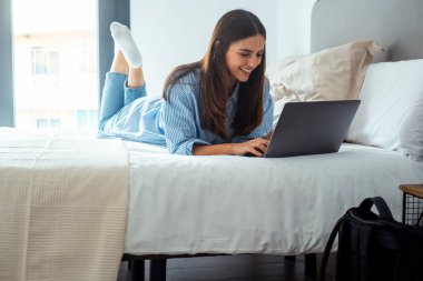 Beyaz gömlekli ve kot pantolonlu genç bir kadın rahat bir dairede dizüstü bilgisayarla çalışıyor..