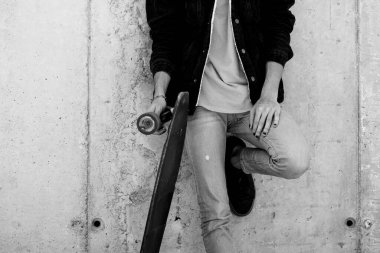 Şehir tarzı giysiler içinde poz veren ve beton duvara karşı uzun paten kayan genç bir gencin tam boy portresi. Siyah-beyaz fotoğrafçılık modası