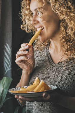 Uzun kıvırcık saçlı, sağlıklı meyve yiyen ve pencereden dışarı bakan kadının yan portresi. Çekici kadın sağlığı iyi doğal yaşam tarzı yer. İçeriyi düşünüyorum. Gülümseyen kadın