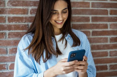 Genç, modern, beyaz kız gülümseyen cep telefonuna bakıyor, tuğla duvara yaslanmış duruyor. Rahatlamış bir kadın telefona bakar. Teknoloji konsepti. Milenyum kadın insanları cep telefonlarını kaydırıyor