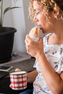  unhealthy woman eating sandwich and working on laptop at home sitting on sofa. Bad nutrition food habits. Gaining weight eating junk food. Fast food lunch during work time indoor clipart