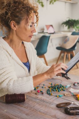 Genç bir kadın evde, online iş aktiviteleri için mücevher ve kolye yapıyor. Evdeki masada oturan kadınlar, tabletle birlikte evde hobi olarak boş vakit geçiriyorlar.