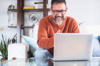 Gözlüklü sakallı adam evde otururken bilgisayarında görüntülü konuşma yapıyor ve gülümsüyor. Cep telefonu kullanan insanlar kavramı..