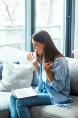 Genç kadın dizüstü bilgisayar kullanıyor, evdeki koltukta oturuyor, güzel kız sosyal ağlarda sohbet ediyor ve arkadaşlarıyla gülüyor. Teknoloji ve arkadaşlık kavramı..