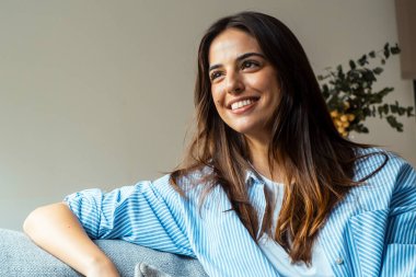 Gülümseyen genç bir kadın rahat bir kanepeye oturur, uzaktan hayal kurar, mutlu bir milenyum kızı rahat bir kanepeye uzanır, oturma odasında dinlenir ve hafta sonunun tadını çıkarmayı düşünür.