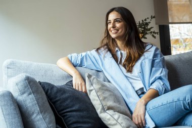 Rahatla ve gevşe. Genç, güzel, esmer, mutlu bir yüzü olan kadın kanepede tek başına gülümsüyor. Evde dinlenen rahat kadın portresi..