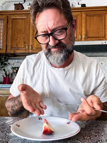 stock image Mature man looking a small piece of fruit in the dish as lunch to eat in dieting weight loss extreme nutrition control. Sad and resigned expression. No carbs diet low calories concept to lose fat