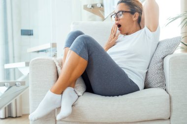 Beyaz sandalyede oturan yorgun kadın spor kıyafetleri giyerek yorgun esniyor. Canlılık ve sağlıklı yaşam tarzı. Kadın insanlar gün ışığında evde uyumaya hazır. Boşluk portresini kopyala tembel