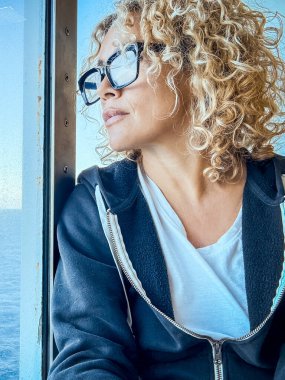 Beautiful woman with glasses spending free time looking at the sea. She takes care of herself, relaxing. Enjoy the view, look through the window. Middle-aged blonde and curly female looks thoughtfully. Peaceful mornings. clipart