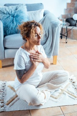 Orta yaşlı bir kadın evde meditasyon yapıyor oturma odasında halının üzerinde oturuyor gözleri kapalı ve vücudu bakımlı. Nefes alma ve rahatlama rutini. Sağlıklı yaşam tarzı, ev hanımları.