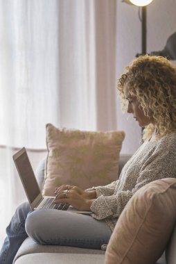 Evde kanepede dizüstü bilgisayar kullanan kıvırcık saçlı sarışın kadın. 45 yaşında, ciddi bir orta yaşlı kadın internette sörf yaparken, web sitesinde alışveriş yaparken ve oturma odasındaki kanepede dinlenirken bilgisayara bakıyor..