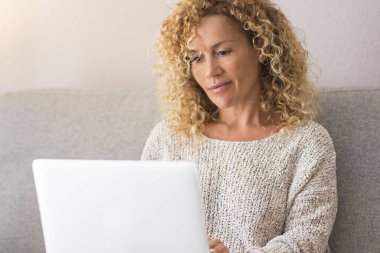 Happy blonde woman with curly hair using laptop sitting on sofa at home. Smiling 45 year old middle aged woman looking at computer while surfing internet, shopping on website and relaxing on living room sofa. clipart