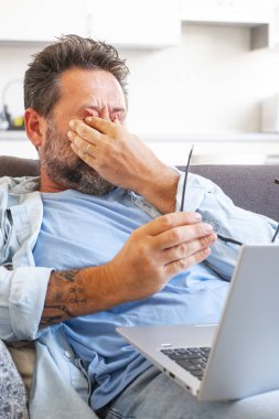 Yorgun işadamı gözlüğünü çıkarıp dizüstü bilgisayarla çalışmaktan gözünün yorulduğunu hisseder, beyaz adam kanepeye oturur ve gözlerini ovuşturur, baş ağrısı ve enerji eksikliği hisseder.