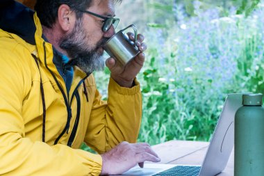 Sakallı gezgin açık havada kahve içiyor. Boş zamanlarında, tahta bir masada oturmuş özgürlüğün ve yolculuğun tadını çıkarıyor. Çevrimiçi olarak her yerde laptop bağlantısı olan modern işçiler. 
