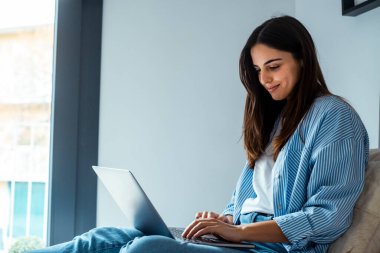 Oturma odasındaki kanepede rahat rahat otururken, genç milenyum kadını laptopuyla evden çalışıyor. İnternette yurtiçi sorunlara çözüm ve günlük yaşamdaki zorluklar için rahat ve modern bir yaklaşımla gezinmek