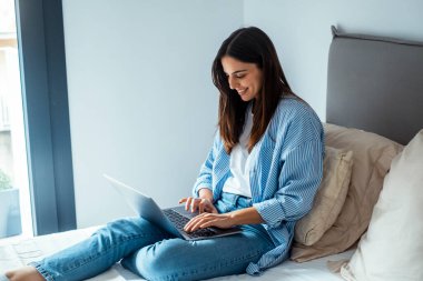 Genç profesyonel bir kadın otel yatağında kablosuz kablosuz kablosuz bağlantısı olan bir dizüstü bilgisayar kullanarak rahatça oturuyor. İş için seyahat eden ve online çalışan dijital göçebelerin modern yaşam tarzını yakalar