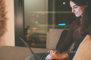 Geç saatlere kadar çalışma: Gülümseyen bir üniversite öğrencisi kanepede oturur, bilgisayarında çalışır ve yararlı sonuçlar için internete göz atar, sınavlara ve gelecekteki kariyerine odaklanmış ve kararlı bir şekilde hazırlanır