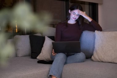 Stresli bir kadın gece geç saatlere kadar bir dizüstü bilgisayarla çalışıyor, baş ağrısı yüzünden alnına dokunuyor, karanlık bir oturma odasındaki bir kanepede tek başına oturuyor, önemli bir iş projesine yetişmeye çalışıyor.