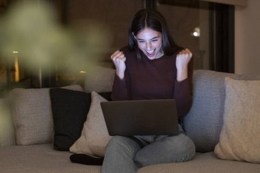 Dizüstü bilgisayarının önünde kutlama yapan mutlu bir kadın, önemli bir dönüm noktasını geçtikten sonra sevinçle seviniyor, örneğin bir spor bahsini kazanmak, sınavı geçmek, yeni bir iş bulmak, ya da online bir randevu, teknolojinin gücünü kucaklamak gibi.