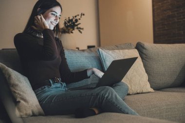 Evdeki kanepede dinlenen gülümseyen bir kadın, internette gezinmek ve gece geç saatlerde web sitelerini okumak için dizüstü bilgisayar kullanıyor. Rahat bir dairede teknolojiyle vakit geçirmek, kablosuz internet ve dijital ortamın tadını çıkarmak.