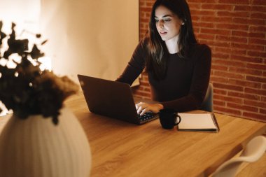 Şirin ve profesyonel bir kadın, akşamları evinde huzur içinde çalışıyor ve gülümsüyor. Dizüstü bilgisayardaki işiyle eğlenirken rahatlıyor, serbest yaşamı kucaklıyor ve modern uzaktan kumandayla çalışıyor