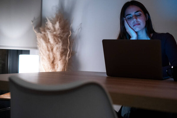Ночной портрет уставшей и измученной женщины, работающей над своим ноутбуком, демонстрирующий скуку и разочарование. Одинокая дома, она борется с бессонницей, глядя в компьютер, не в состоянии заснуть из-за чрезмерной интернет-работы.