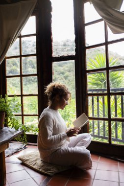 Huzurlu bir rahatlama ve farkındalık anı. Bir kadın evde tek başına oturmuş kitap okurken. Etrafı pencerelerden görülebilen doğa ile çevrili kadın, akıl ve beden için sağlıklı bir yaşam tarzını benimsiyor.