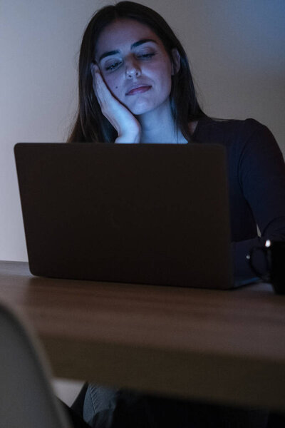 Ночной портрет уставшей и измученной женщины, работающей над своим ноутбуком, демонстрирующий скуку и разочарование. Одинокая дома, она борется с бессонницей, глядя в компьютер, не в состоянии заснуть из-за чрезмерной интернет-работы.
