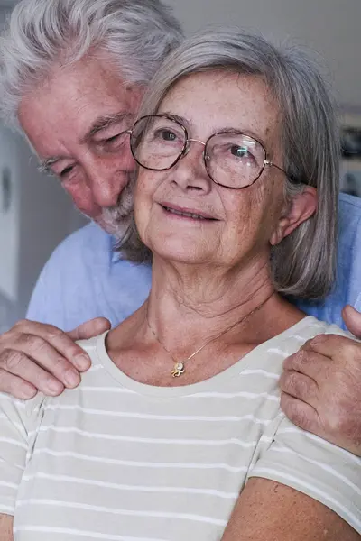 stock image Front view of a mature couple in their home, an elderly man embracing his wife from behind with love and protection. The romantic grandparents display affection and good health, enjoying their time together