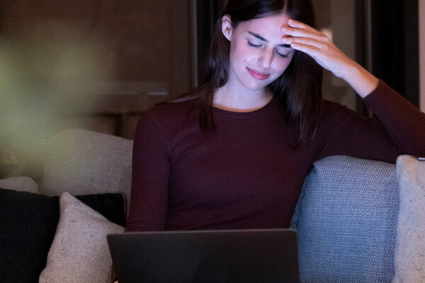 Stressed woman working late at night on a laptop, touching her forehead due to a headache, sitting alone on a sofa in a dark living room, struggling to meet an important work project deadline
