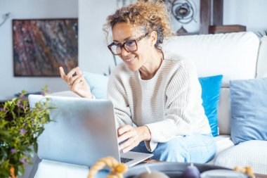 Evdeki oturma odasındaki kanepede dizüstü bilgisayar kullanarak video aramakla meşgul neşeli bir bayan. İnternette gülümseyen ve iş arkadaşlarıyla konuşan bir kadın. Uzak çalışma bağlantısı