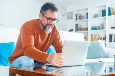 Konsantre yetişkin erkek, evdeki kanepede dizüstü bilgisayarını kullanarak alternatif ofis ayarlarını yönetirken ya da bir sonraki tatil kaçamağını planlarken.