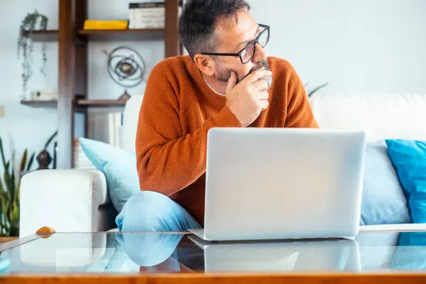 stock image Thoughtful man focused on his laptop while sitting on the sofa at home, managing an online business or conducting research for effective household financial management and vacation planning