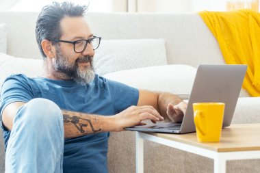 Yetişkin bir adam bilgisayarında uzaktan çalışıyor, rahat bir şekilde kanepede oturuyor, internette geziniyor ve alternatif ofis ortamında bilgi aramak için kablosuz internet kullanıyor.