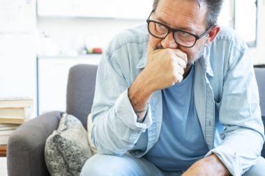 Middle-aged man sitting alone on his couch at home, lost in thought with a serious expression, holding his chin while facing personal struggles related to job loss and financial concerns clipart